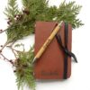 Image shows a leather journal and personalized pen on greenery and berries for the holidays